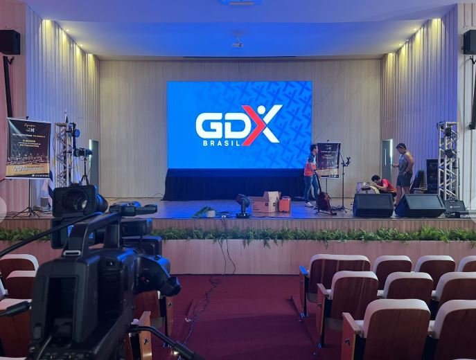 Auditório preparado pela GDX Brasil com grande painel de LED exibindo o logo GDX no palco, câmera de transmissão em primeiro plano e caixas de som instaladas.