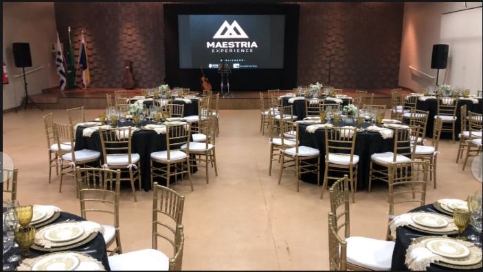 Salão de eventos com mesas redondas e cadeiras douradas voltadas para um palco com telão exibindo “Maestria Experience”, estrutura montada pela GDX Brasil.