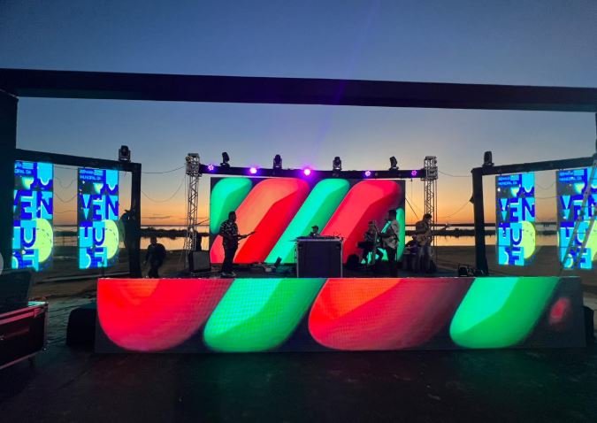 Palco de show ao pôr do sol à beira d’água com painéis de LED coloridos, banda tocando e iluminação de palco montada pela GDX Brasil.